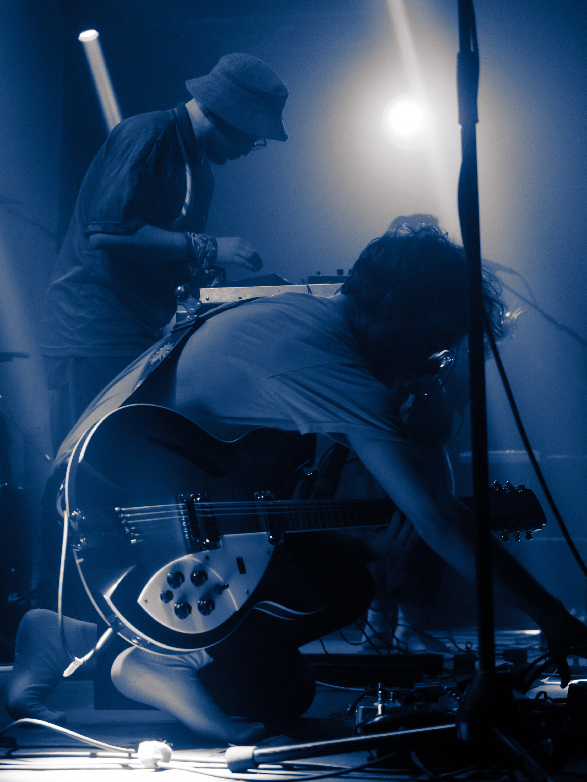 Trevor kneels in the foreground adjusting pedals while Will fiddles with his own pedal board behind him.