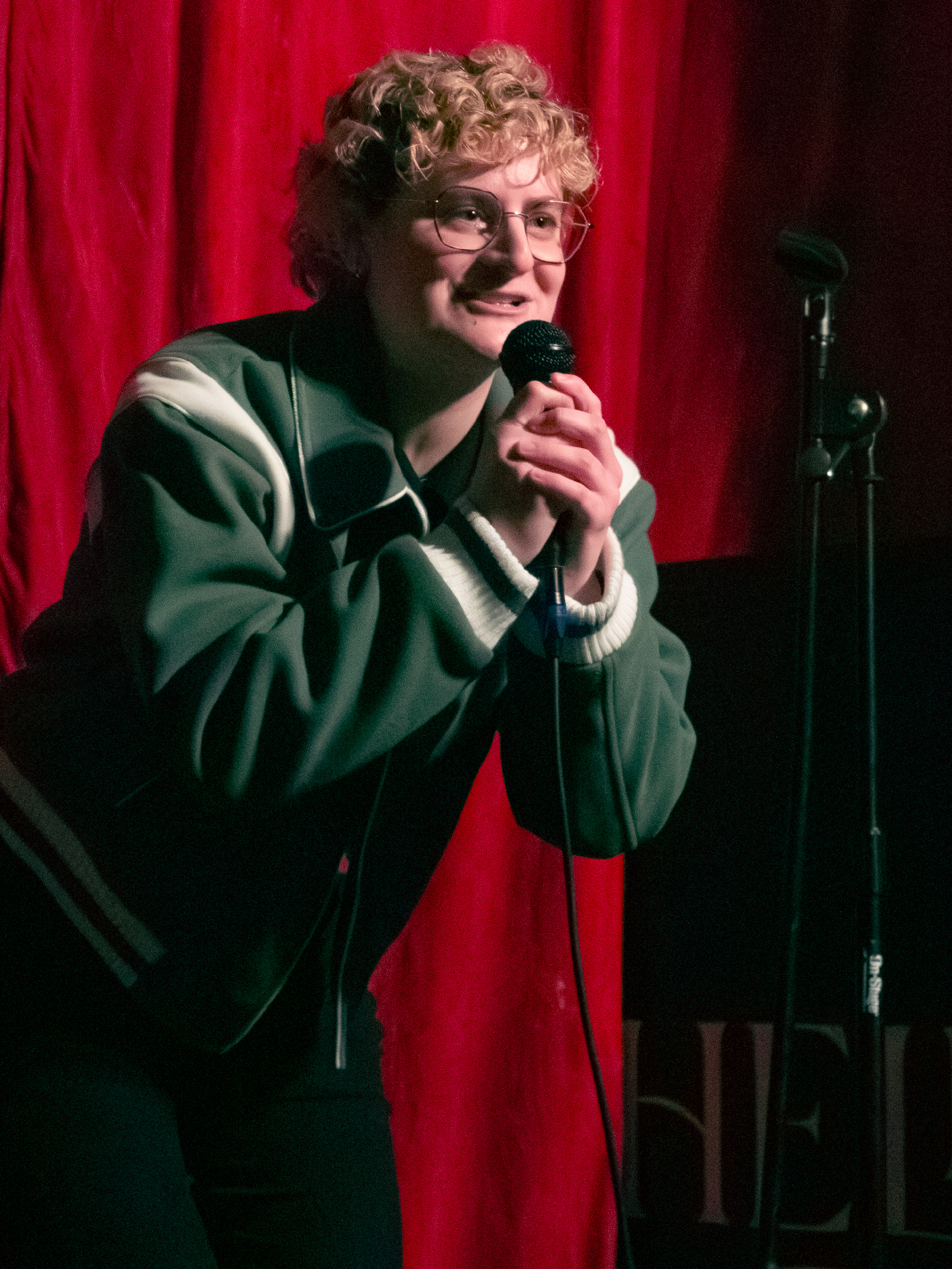 Erica Green tells a joke with both hands on the mic while leaning towards the audience.